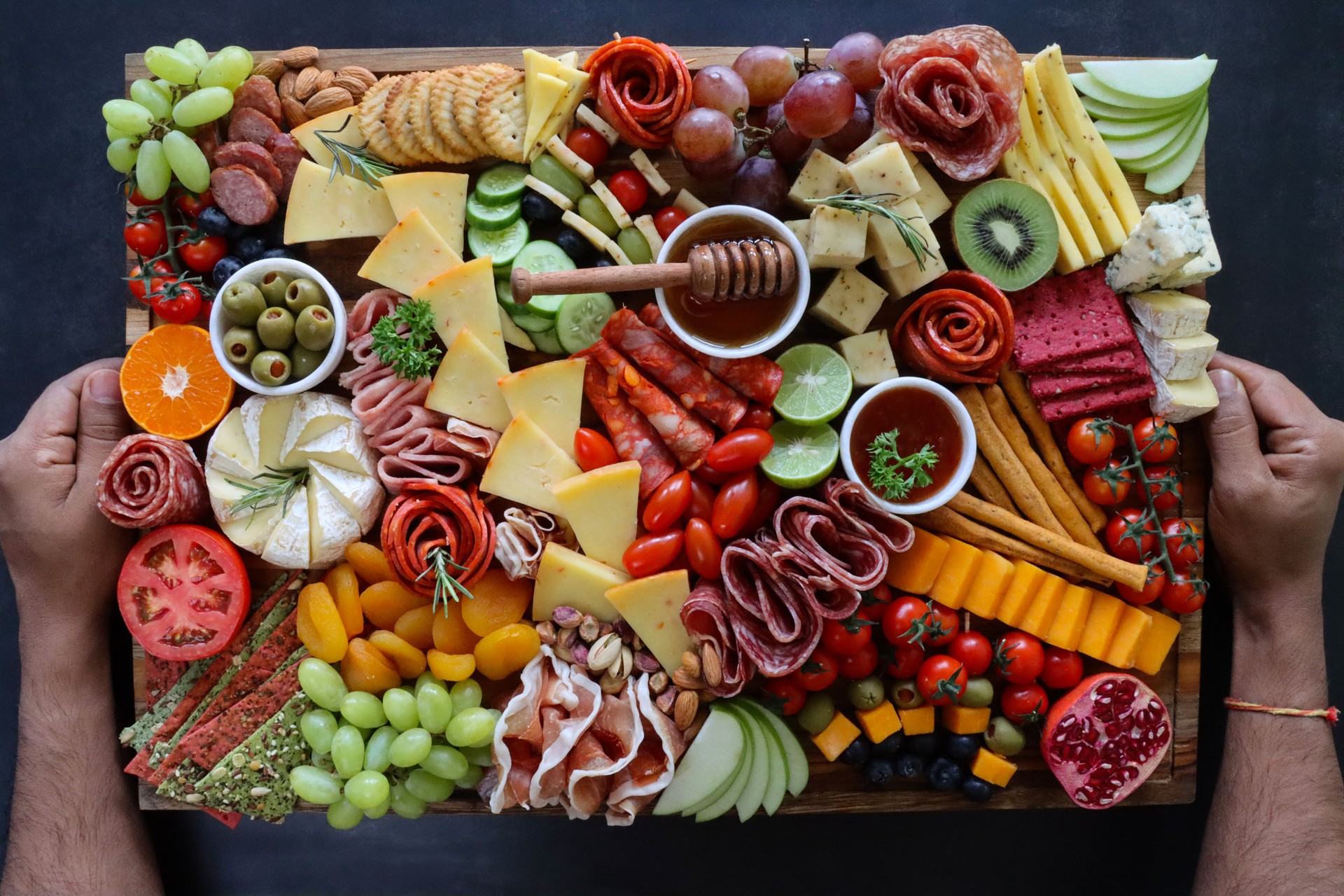 Close-up image of unrecognisable person placing charcuterie board of ingredients on black background, ramekin of honey and dipper, crackers, ham, prosciutto and salami roses, cucumber, lime half, cherry tomatoes, Brie, kiwi, Cheddar, red grapes, elevated