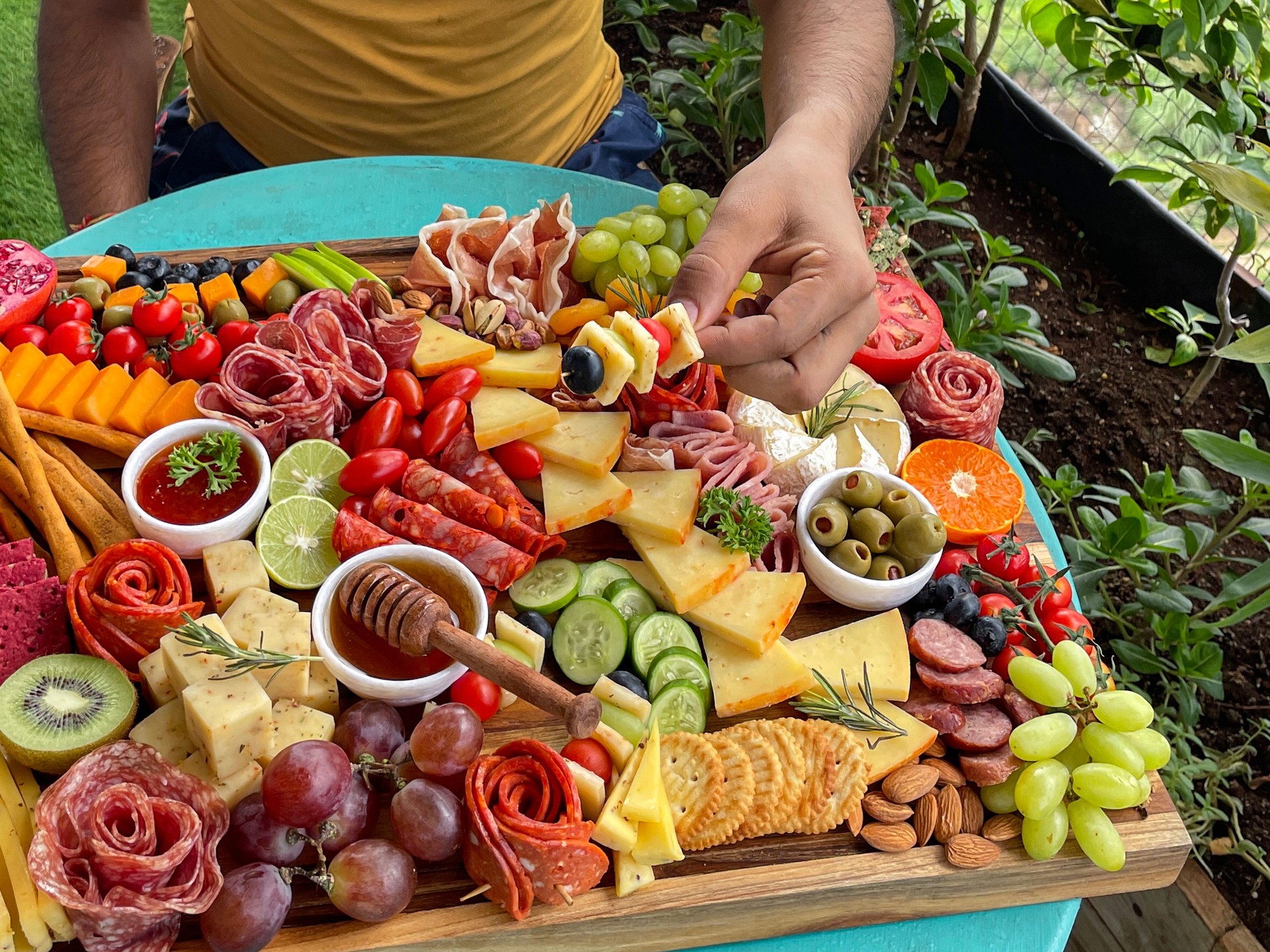 Full frame image of man sat in front of charcuterie board on apartment balcony table, ramekins of honey and chutney, crackers, salami roses, breadsticks, kiwi, vines tomatoes, soft and hard cheeses, stuffed olives, blueberries, elevated view